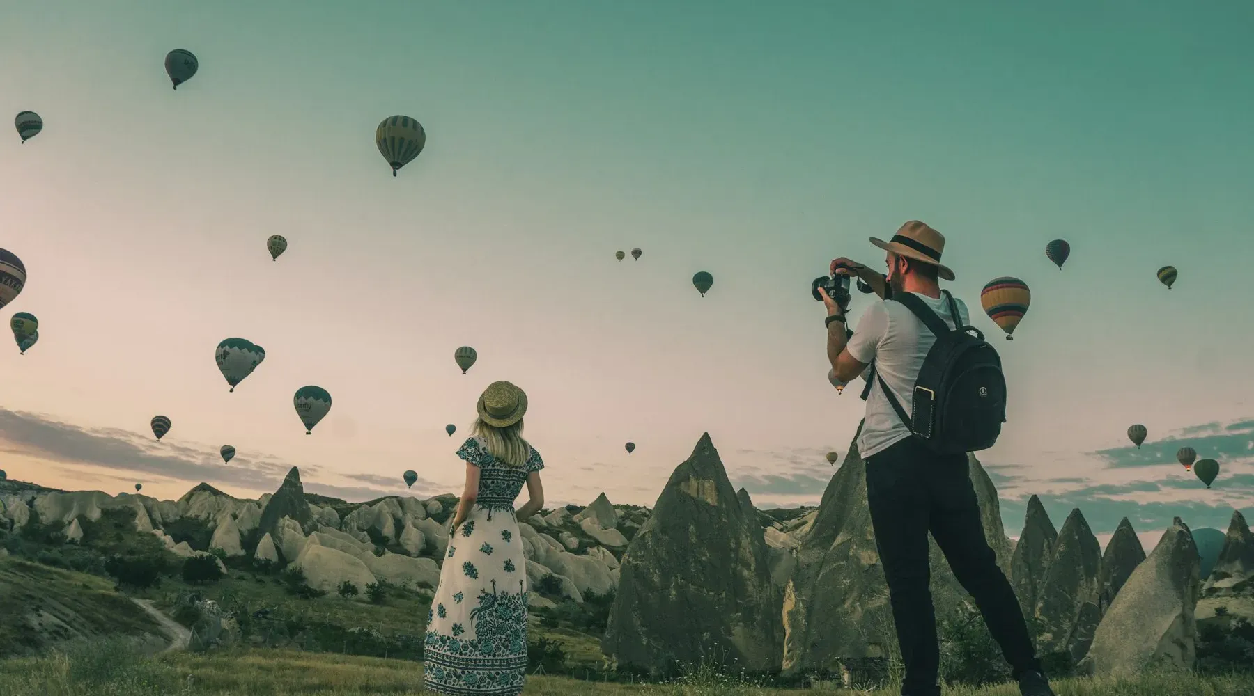 Two travelers photographing hot air balloons over Cappadocia at dawn