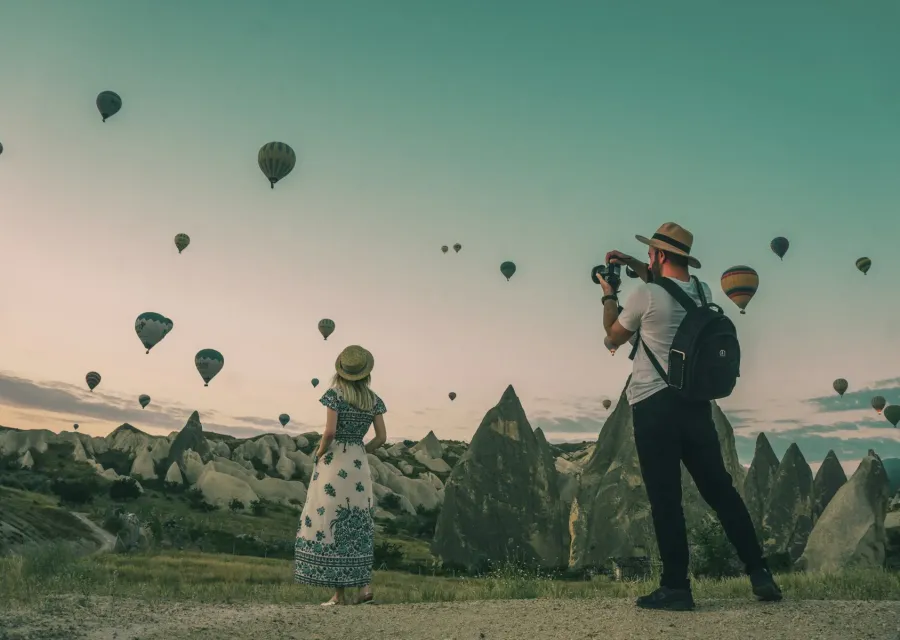 Two travelers photographing hot air balloons over Cappadocia at dawn