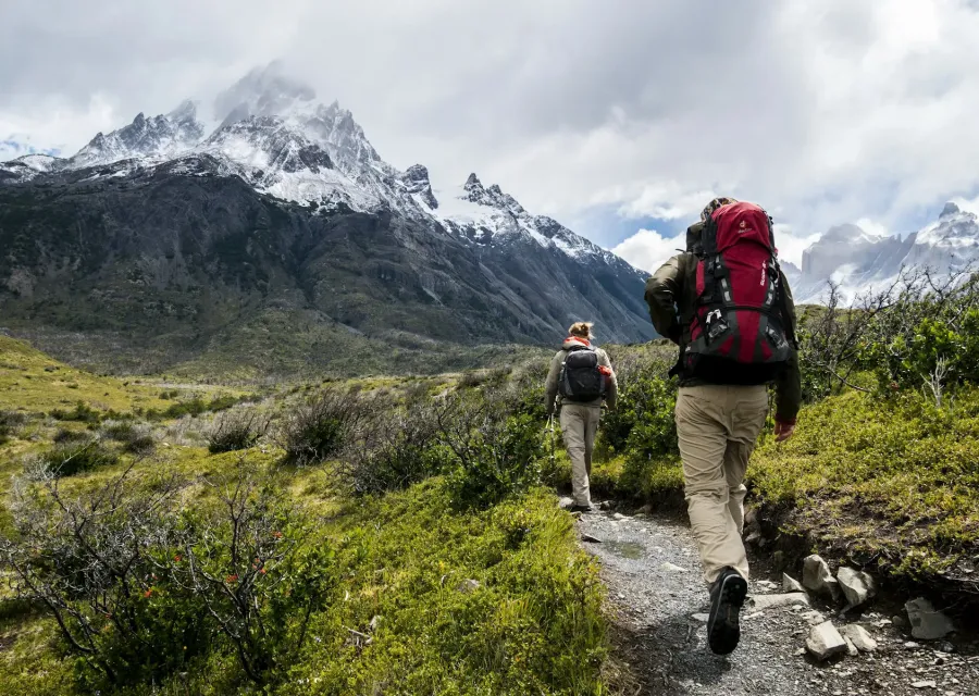 Two hikers with backpacks on a trail below snowy peaks
